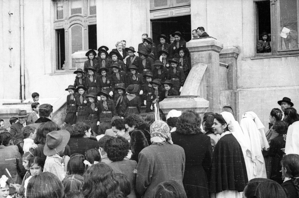 plano general de un grupo de niñas scout paradas sobre unas escaleras vistiendo su uniforme. Hay un grupo de Hombres, Mujeres, Niñas y Niños observándolas en las afueras de la Municipalidad de La Unión