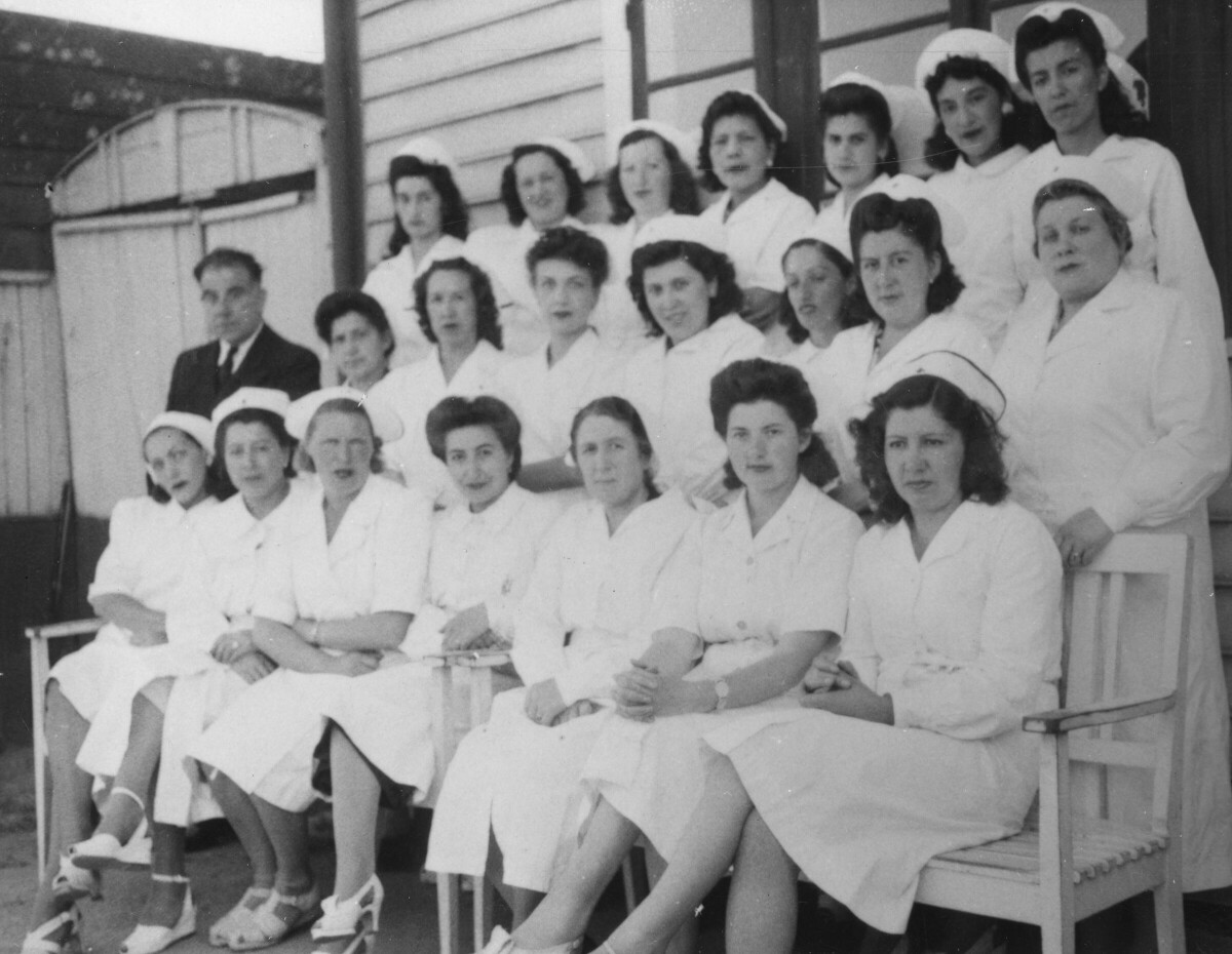Retrato grupal de plano entero de mujeres de la cruz roja, formadas junto a un hombre vestido de Traje. La tercera mujer sentada de izquierda a derecha es Olga Boettcher. Llevan puesto sus uniformes y se encuentran reunidas en las afueras de una estructura no identificada.