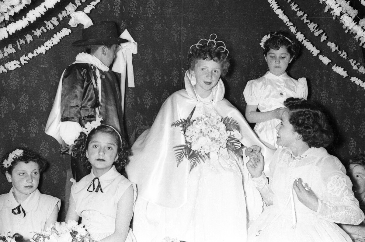Plano entero de un grupo de Niñas y un niño sentados alrededor de una Niña coronada a reina. Se encuentran sentados sobre una tarima decorada por un telón y guirnaldas de flores. Se encuentran en el interior del gimnasio de la Escuela superior de niñas Nº2 durante una actividad escolar