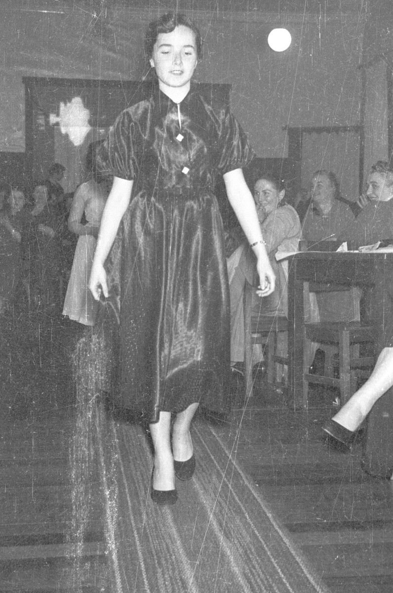 Plano entero de un Mujer Joven modelando un Vestido mientras camina por una alfombra. detrás de ella le sigue otra mujer, modelando también. En un costado y en el fondo hay un grupo de mujeres que las observan, las cuales están sentadas alrededor de unas mesas en el interior de un loca.