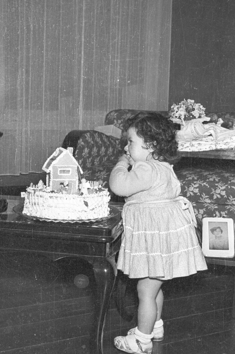 Plano entero de un niña parada con una mano en la boca. Lleva puesto un Vestido y está junto a una mesa sobre la cual hay una torta encima, la cual tiene una casa pequeña y figuras de decoración. Está en el interior del living de una casa