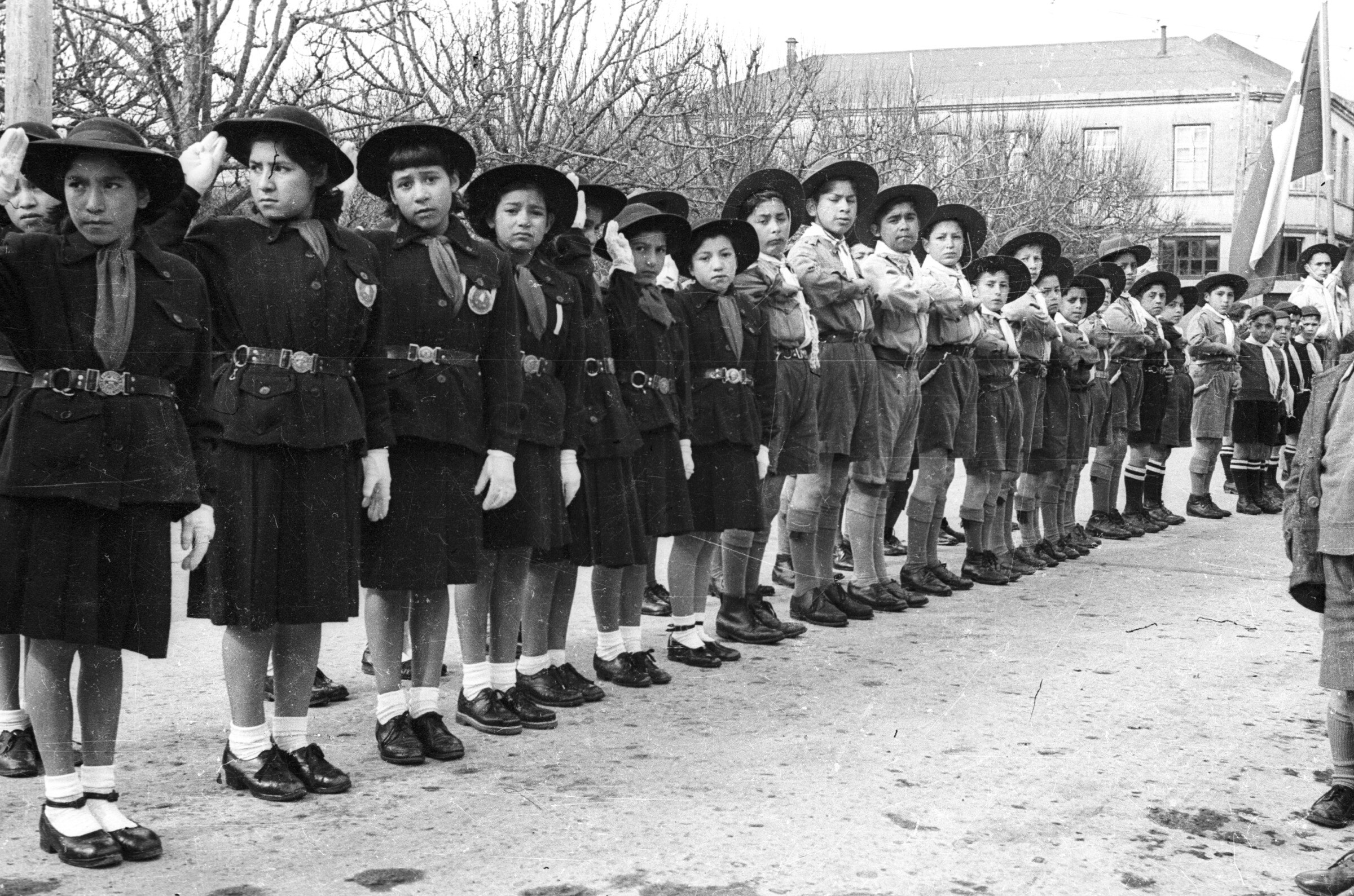 Plano entero de un grupo de Niñas y Niños formados en una hilera mientras realizan un saludo hacia su costado derecho. Todos son pertenecientes a la brigada de scout y llevan puesto su uniforme de ceremonias. Al final de la hilera, hay un hombre parado sosteniendo por el mástil, una bandera chilena. Están ubicados en un costado de la plaza de La Concordia durante un desfile.