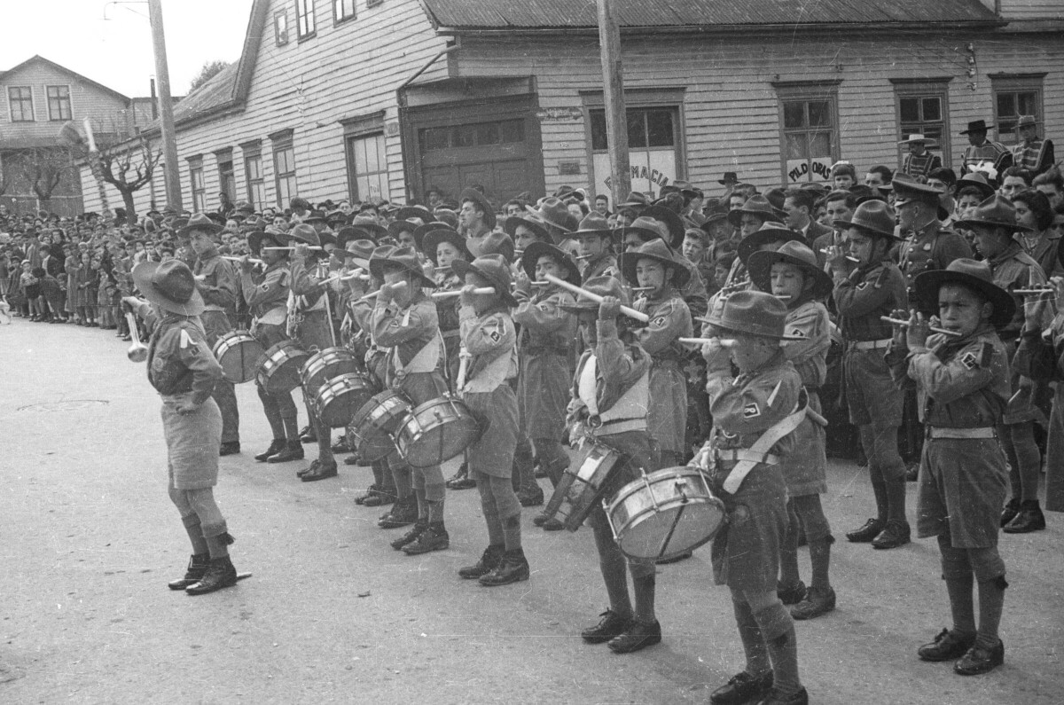 Plano entero de la orquesta sinfónica de la brigada de scout infantil realizando una presentación a vistas de un grupo de Hombres, Mujer, Niñas y Niños ubicados a un costado de la calle. Los Niños se encuentran formados en hileras mientras tocan sus instrumentos en el desfile realizado en la ciudad de La Unión