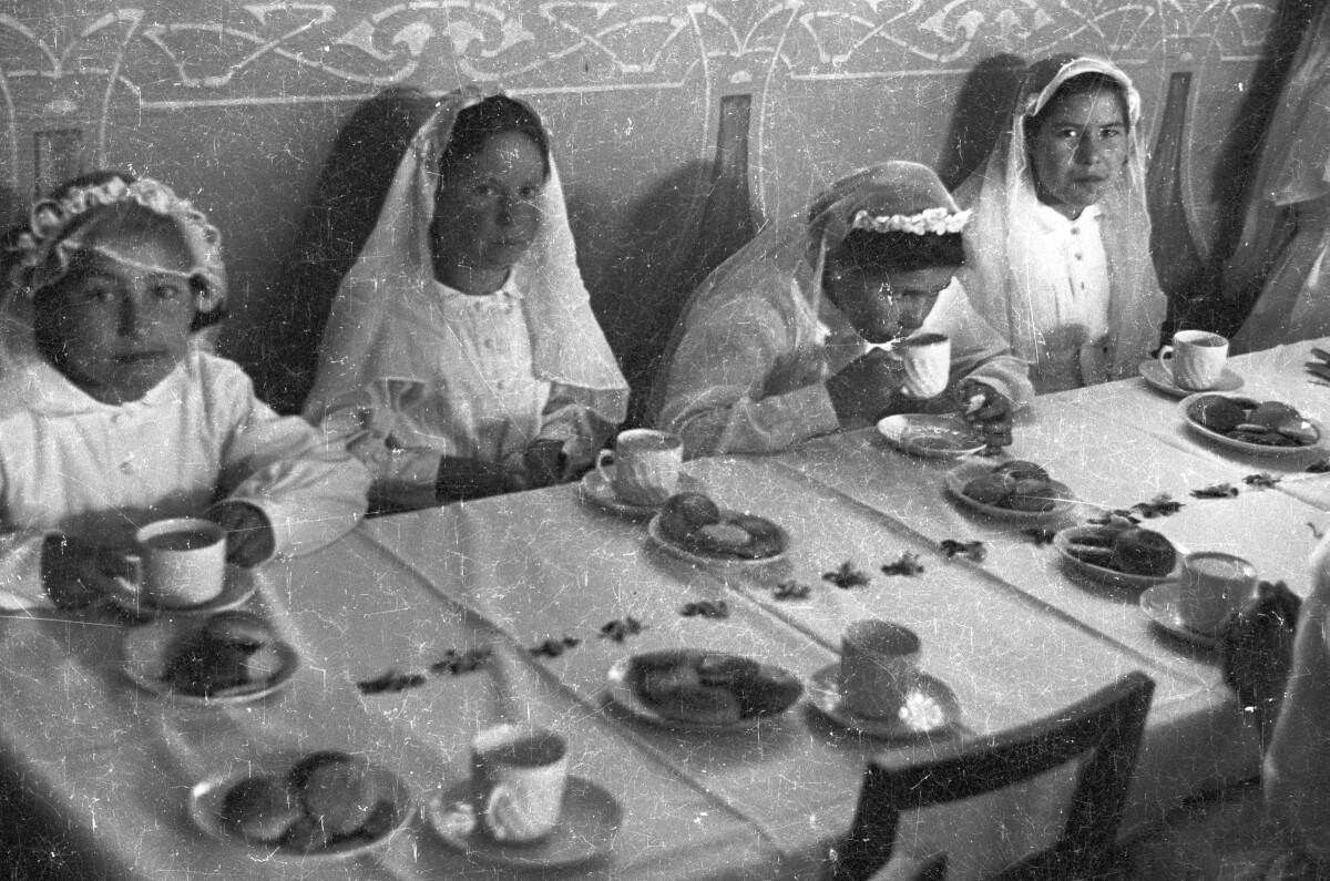 Plano entero de tres niñas mirando hacia la cámara mientras la cuarta está bebiendo de una taza. Se encuentran sentadas alrededor de una mesa. Están en el interior de una infraestructura no identificada celebrando su primera comunión