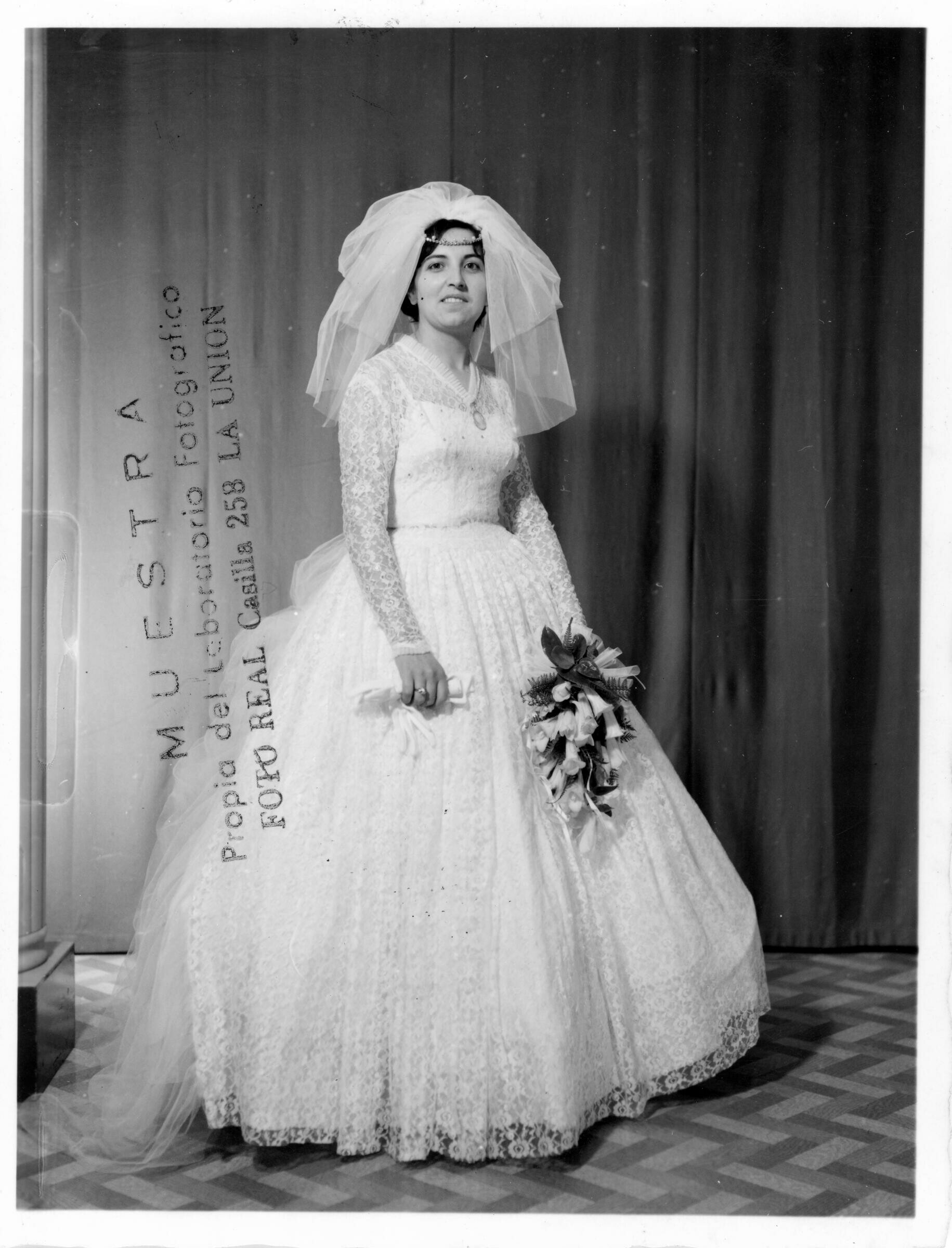 Retrato de estudio de Plano entero de Sylvia García vestida de novia, sonriendo con un ramo de copihues blancos en la mano y sus guantes en la otra.