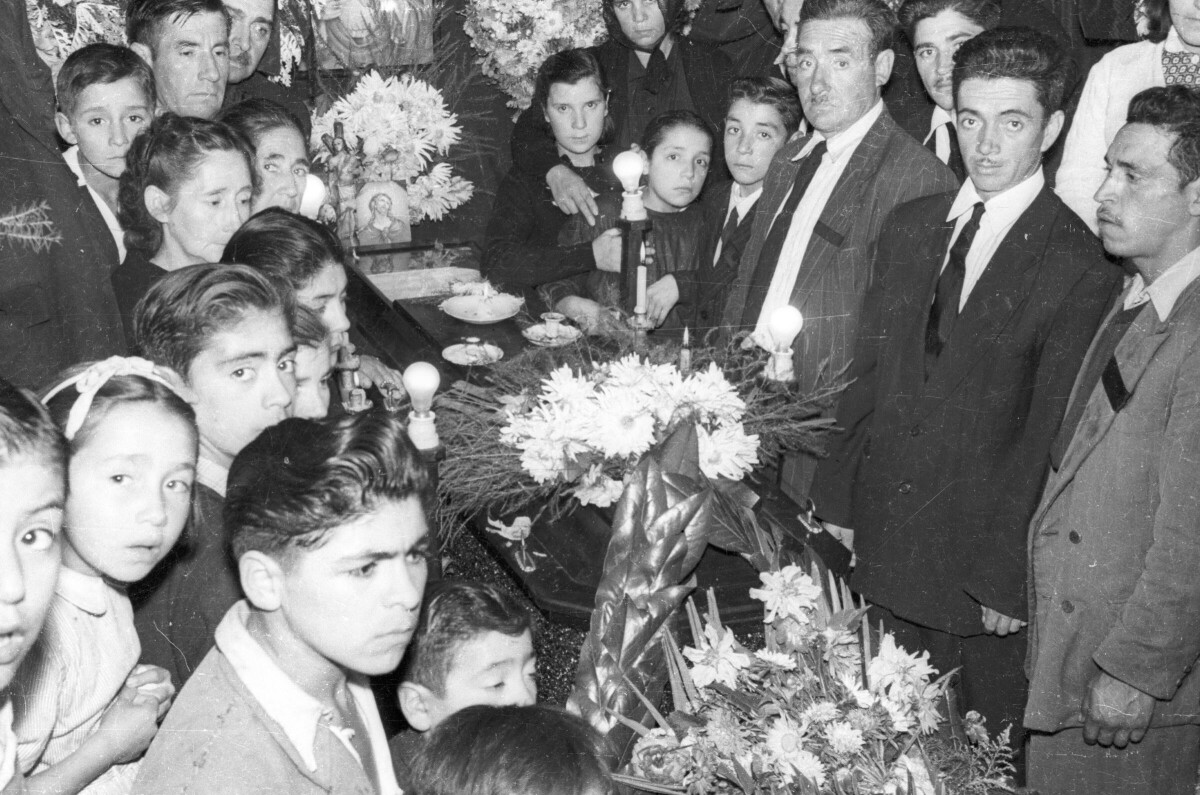 plano general de un grupo numeroso de Mujeres, Hombres, Niñas y Niños mirando hacia la cámara mientras se encuentran parados alrededor de un ataúd, el cual está decorado con coronas de flores e imágenes religiosas. Se encuentran en el interior de una infraestructura no identificada durante un velorio