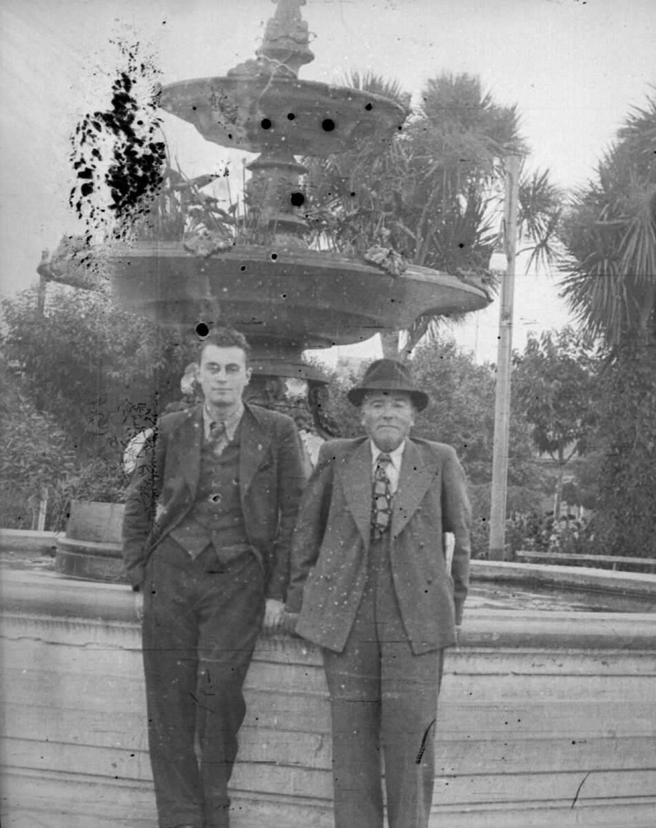 Plano entero de dos hombres, uno joven y otro mayor, posando de pie frente a una fuente de agua en una plaza