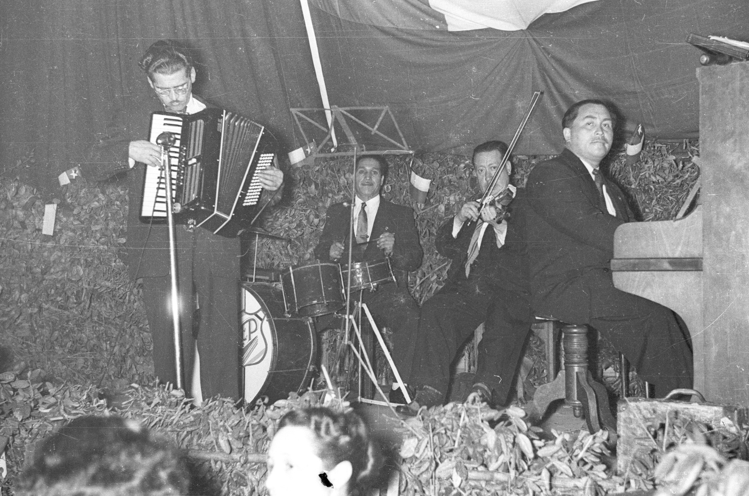 Plano entero de un grupo musical no identificado, realizando su presentación sobre un escenario. Uno de los hombres se encuentra tocando un acordeón, de pie junto al micrófono. Están en el interior de un local durante una celebración de fiestas patrias.