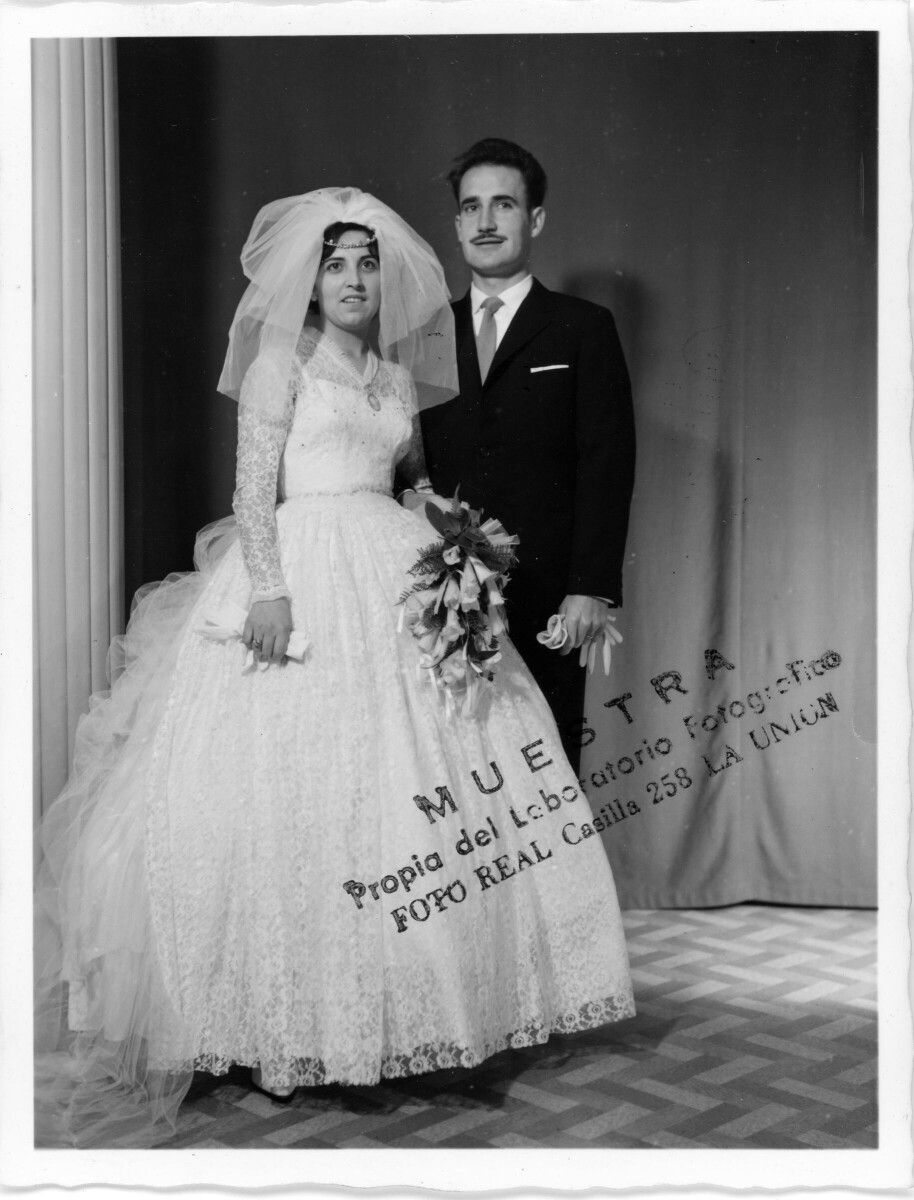 Retrato de estudio de Plano entero de los novios Rudy Pinuer y Sylvia Garcia tomados de las manos. La novia lleva consigo un ramo de copihues blancos. Ambos sonríen y se encuentran parados junto a un pilar.