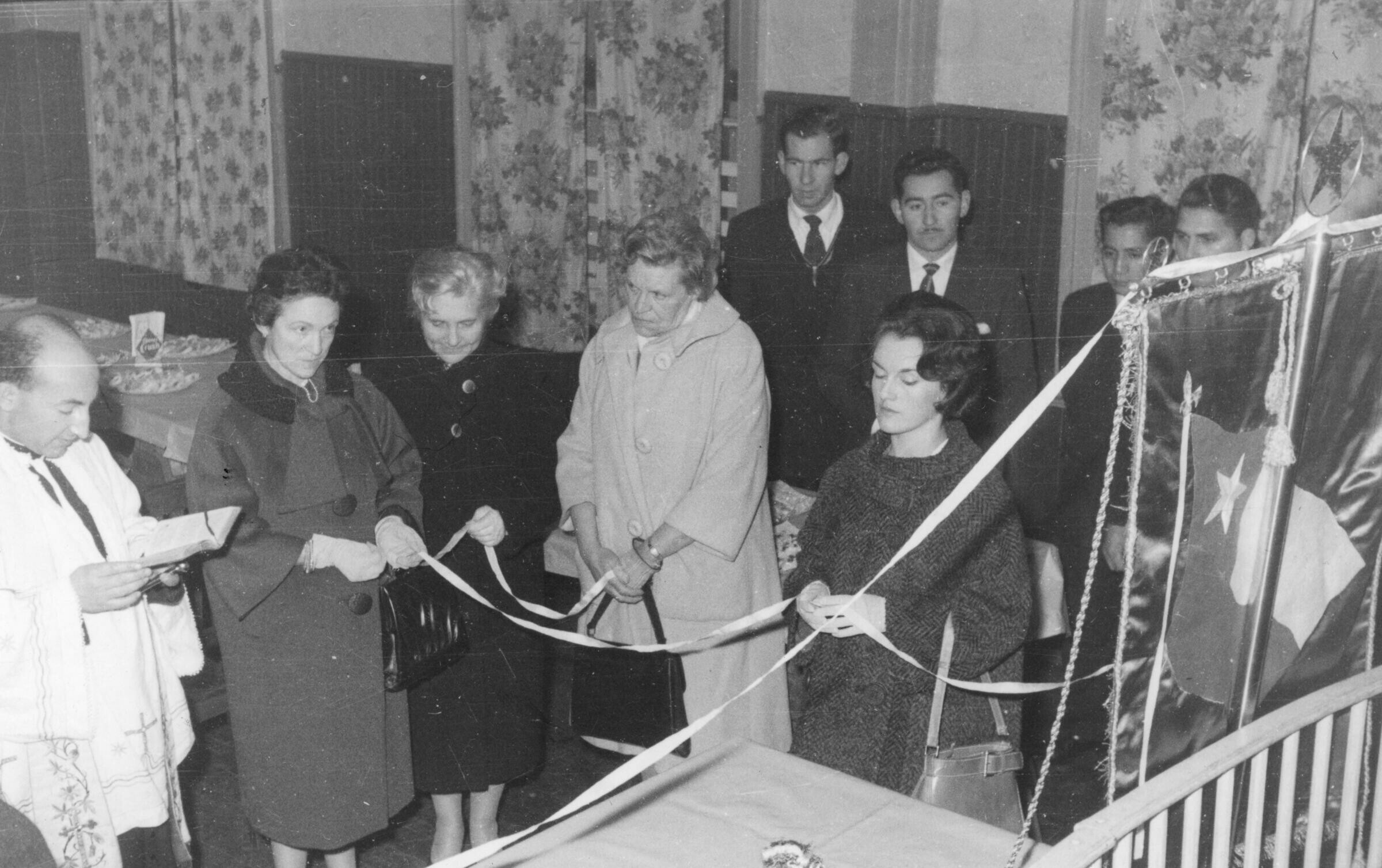 plano general de un sacerdote leyendo la bíblica junto a un grupo de mujeres quienes están sosteniendo las cintas que vienen desde un pendón con la bandera chilena. La tercera mujer de izquierda a derecha es Olga Boettcher. más en el fondo, hay un grupo de hombres vestidos de Traje parados. Se encuentran en el interior de lugar no identificado