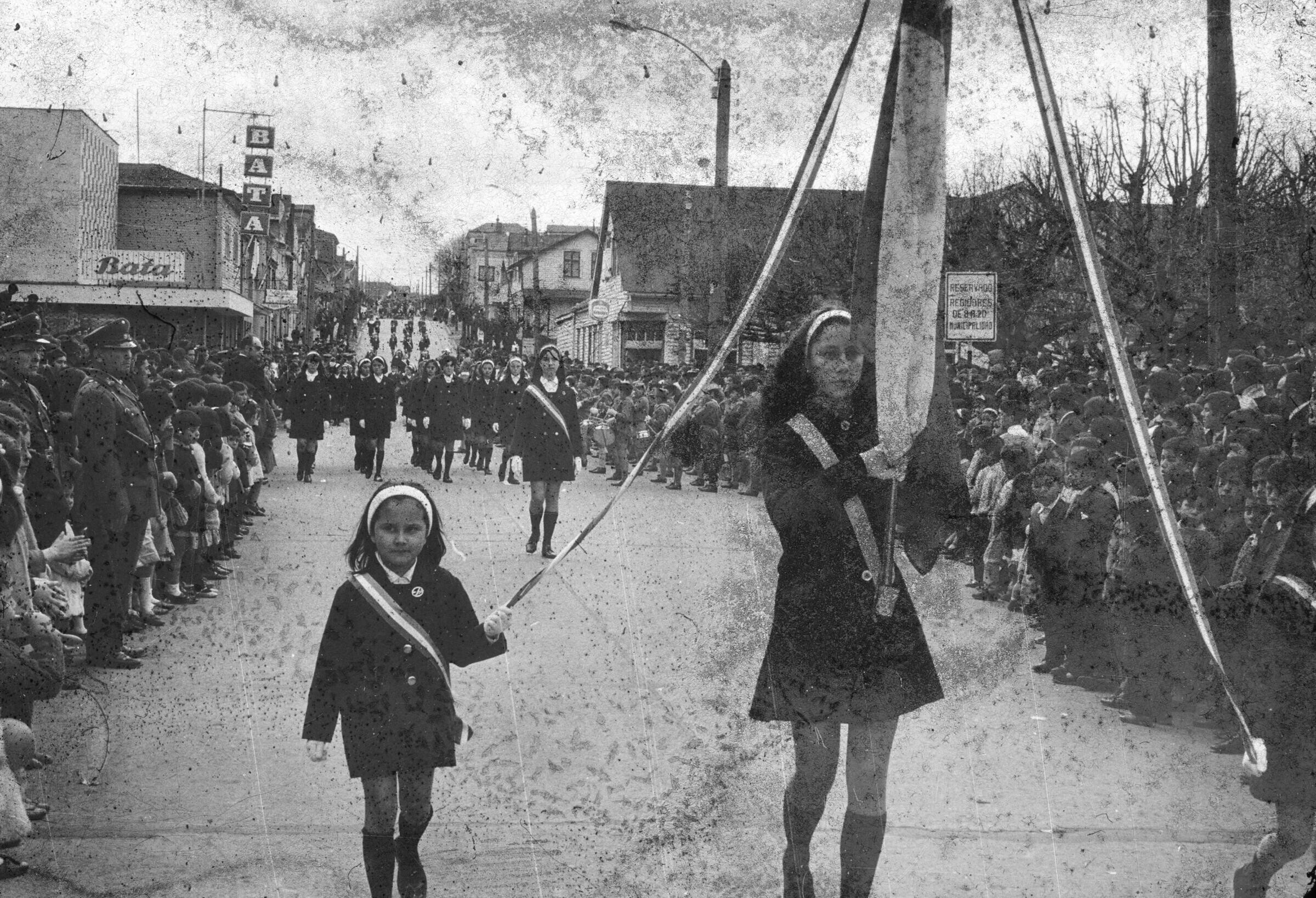 plano general de tres niñas desfilando mientras sostienen las cintas de una bandera. A su alrededor se observa a un grupo numeroso de personas paradas a un costado de la plaza de la Concordia