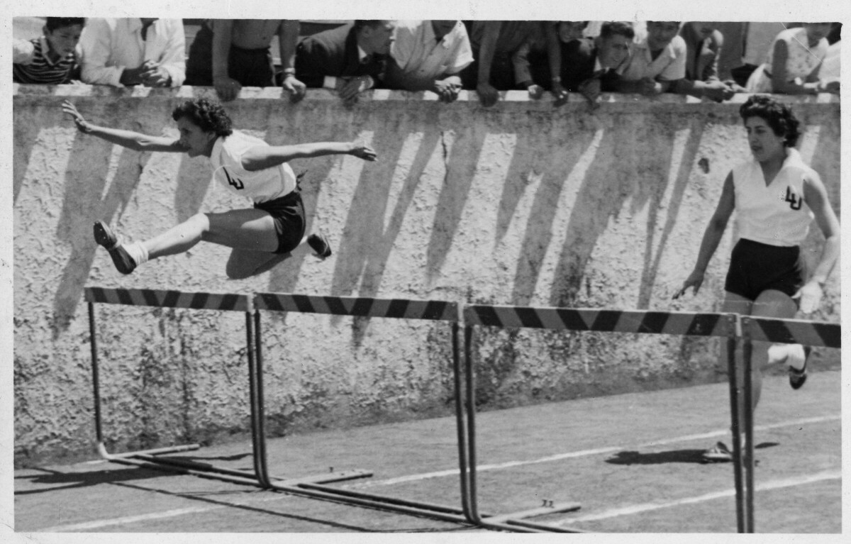 -plano general de una mujer en el aire, saltando una valla. Por detrás otra mujer avanza sobre la pista, Ambas llevan bordado en sus poleras las iniciales --LU--. A un costado, un grupo de hombres les animan desde las graderías. Se encuentran en un zonal sur de atletismo realizado en Tomé.-
