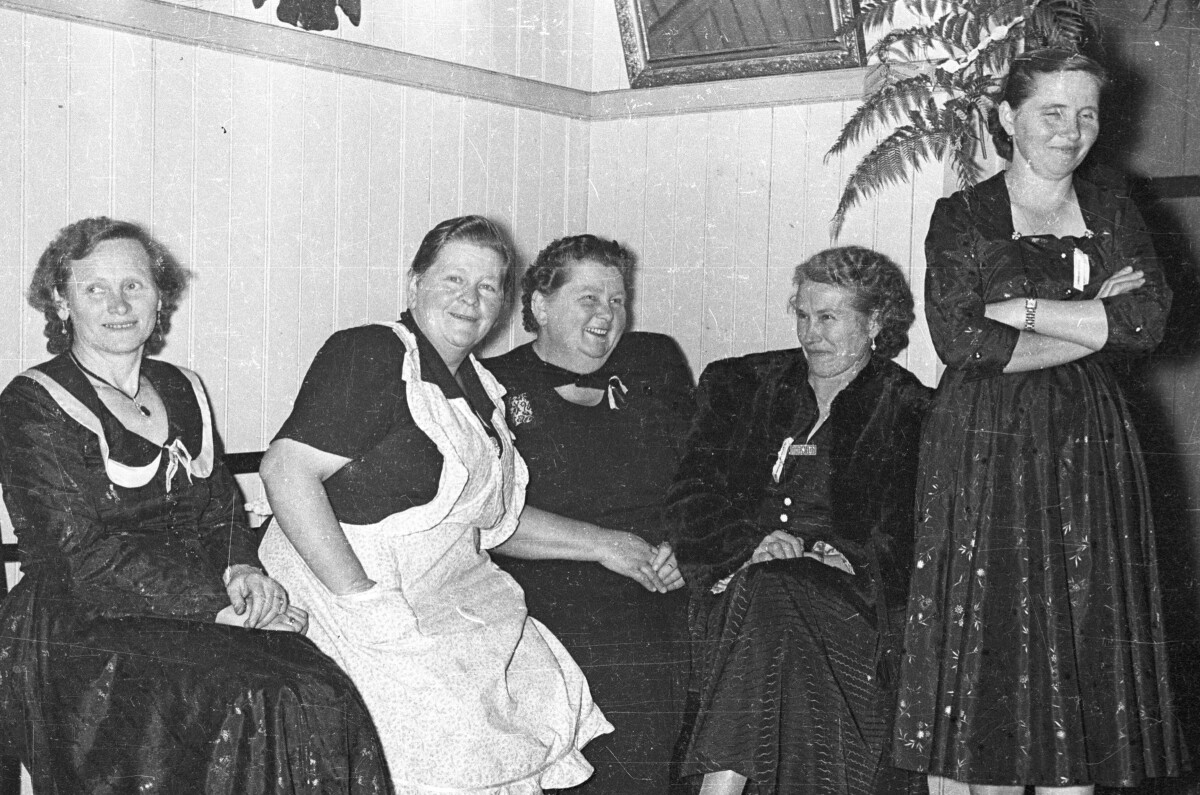 Plano entero de un grupo de mujeres, riendo mientras se encuentran sentadas junto a una pared. Una de ellas, está parada con los brazos cruzados, mientras sonríe. Se encuentran en el interior de una casa durante la celebración de una fiesta de bodas