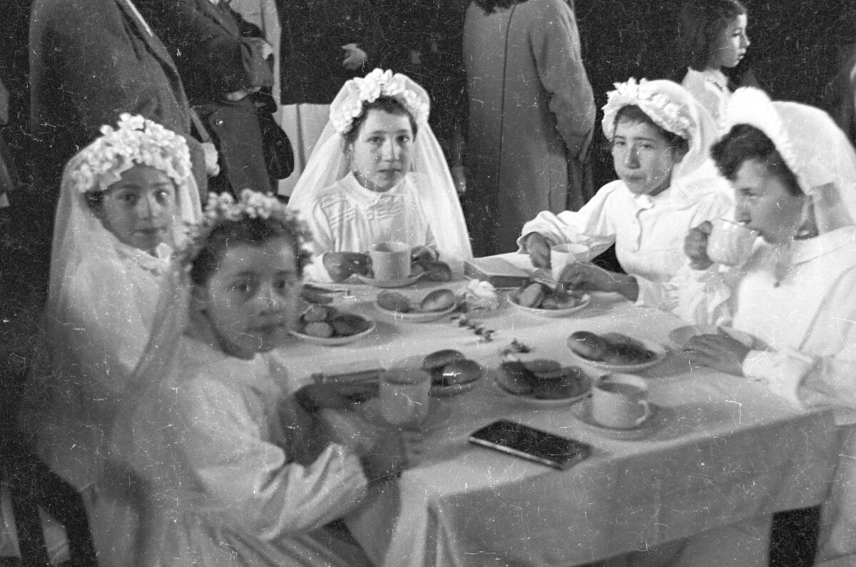 Plano entero de un grupo de Niñas mirando hacia la cámara. Se encuentran sentadas alrededor de una mesa, tomando el té. A su alrededor un grupo de personas va caminando por un costado. Están en el interior de una infraestructura no identificada celebrando su primera comunión