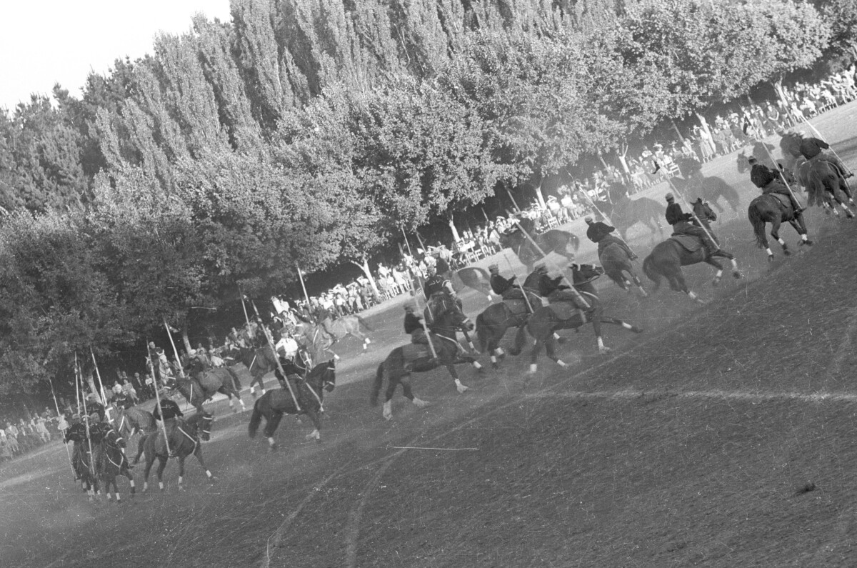 plano general de un grupo de jinetes a caballo, cabalgando en un circulo mientras llevan una valla en sus manos realizando una presentación. En el fondo, Niñas, Hombres y Mujer los observan sentados a un costado de la cancha ubicada en el estadio Carlos Vogel