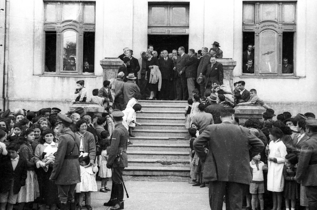 plano general de un grupo numeroso de personas reunidas en la vereda mientras que Carlos Vogel y un grupo de Hombres se encuentran conversando en el frontis de la municipalidad
