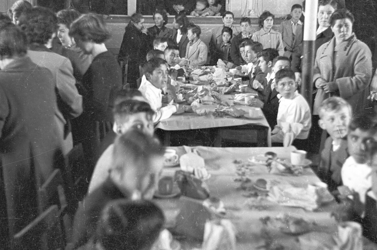 plano general de un grupo de Niños sentados alrededor de dos mesas, sobre las cuales hay galletas, pan y tazas de té. A su alrededor, un grupo de mujeres se encuentran conversando. Están en el interior de una infraestructura no identificada celebrando su primera comunión
