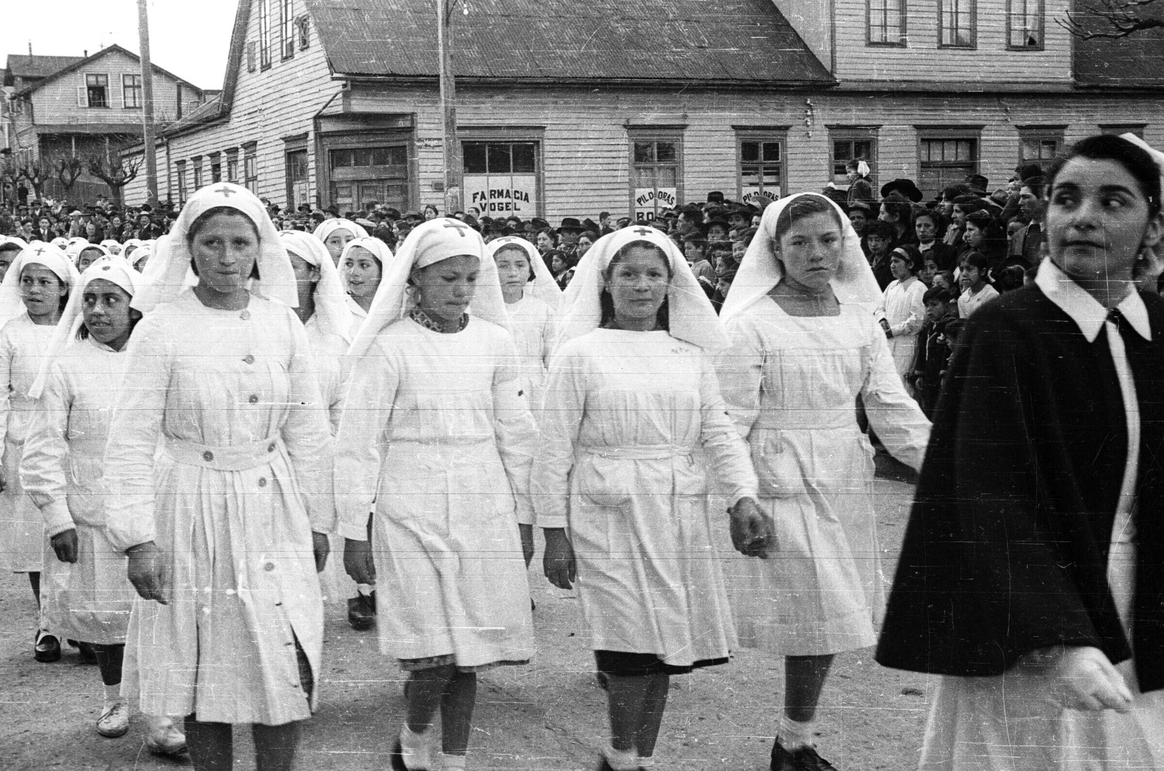 Plano entero de un grupo de Niñas pertenecientes a la Cruz roja juvenil de la escuela Nº2, caminando vestidas de blanco y con un velo en la cabeza por el medio de una calle durante un desfile