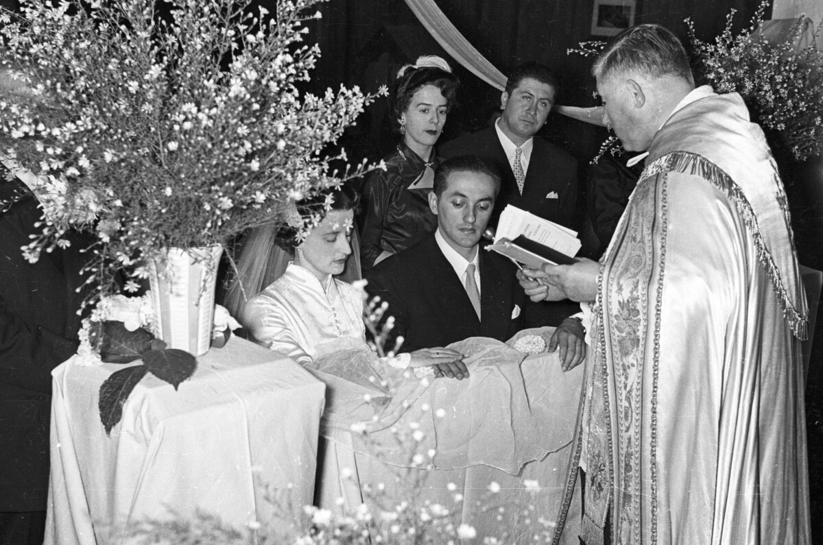 Plano medio largo de un sacerdote leyendo la biblia frente a una pareja de novios que se encuentran hincados en un reclinatorio. Los acompañan en el fondo, sus padrinos de bodas. Se encuentran en el interior de la parroquia San José durante la misa de celebración de su casamiento