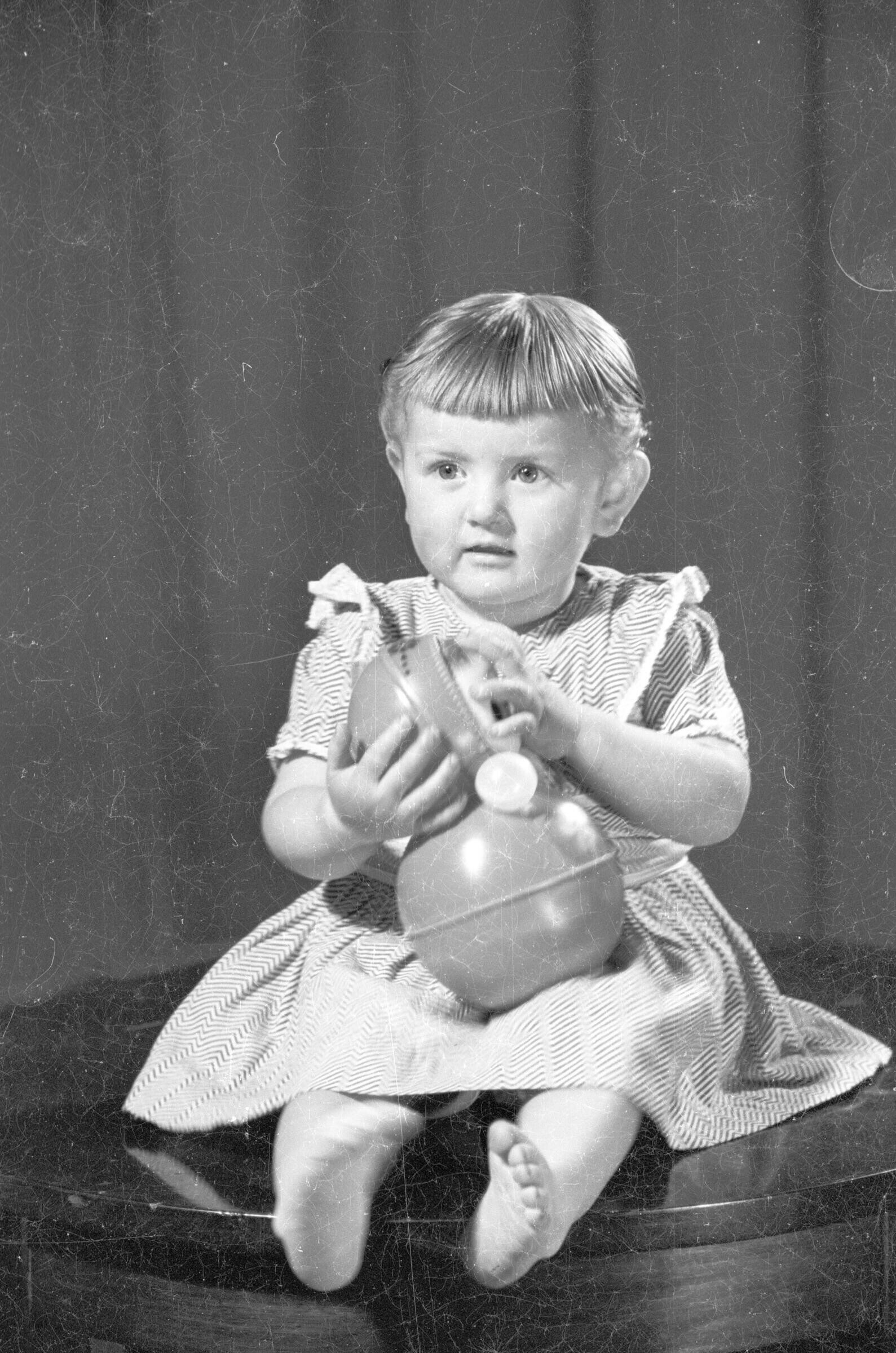 Retrato de estudio de plano entero de una niña con vestido, posando descalza sobre una mesa circular con una muñeca entre las manos