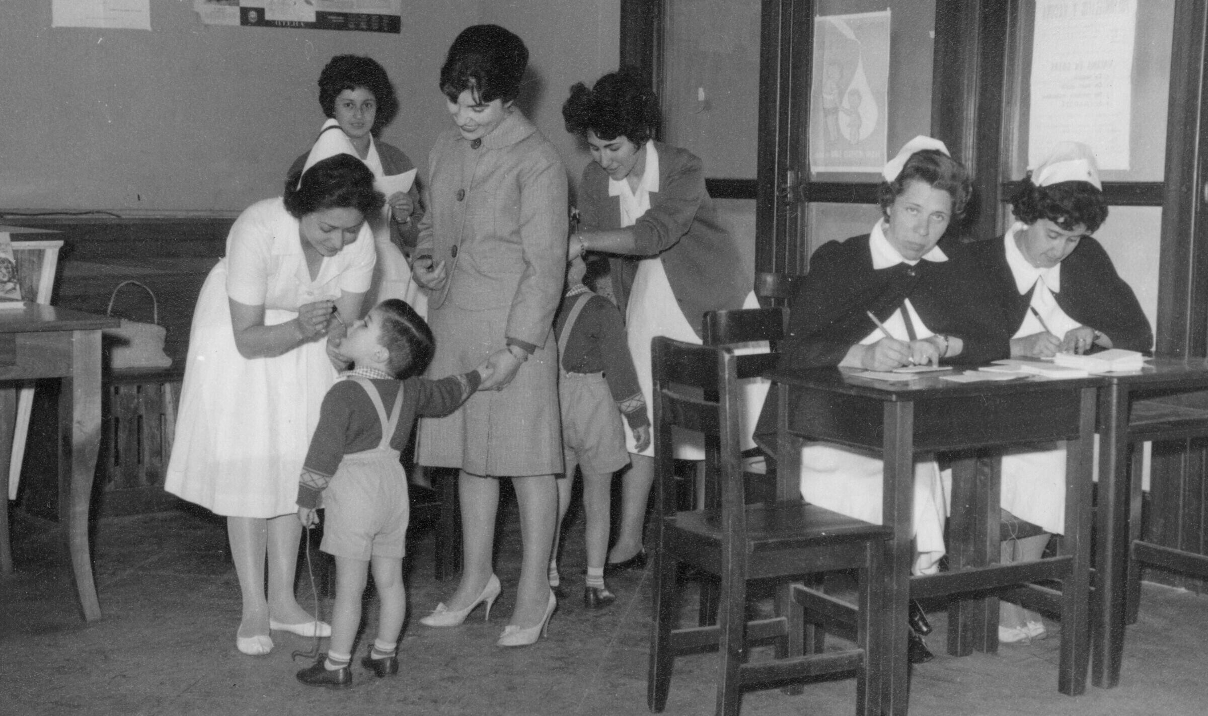 plano general de un voluntaria de la cruz roja, dándole gotitas en la boca a un Niño que se encuentra tomado de la mano de su mamá. detrás de ellas, otra voluntaria repite el procedimiento con otro niño. En un costado, hay otras dos miembros trabajando junto a sus escritorios. Se encuentran en el interior de una infraestructura no identificada.