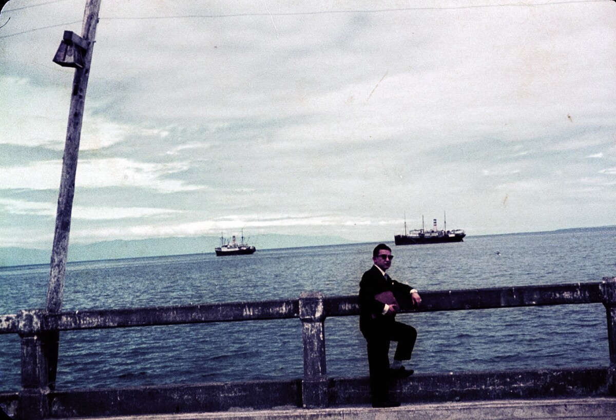 Retrato de plano entero de Hombre con Traje apoyado de una baranda a orilla del mar, con barcos navegando