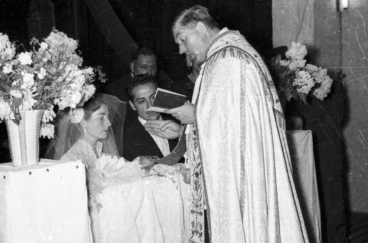 Plano medio largo de un sacerdote leyendo de un libro mientras bendice las argollas de los novios, quienes se encuentran hincados en un reclinatorio. Está llevando a cabo la misa de celebración de su matrimonio en el interior de la parroquia San José