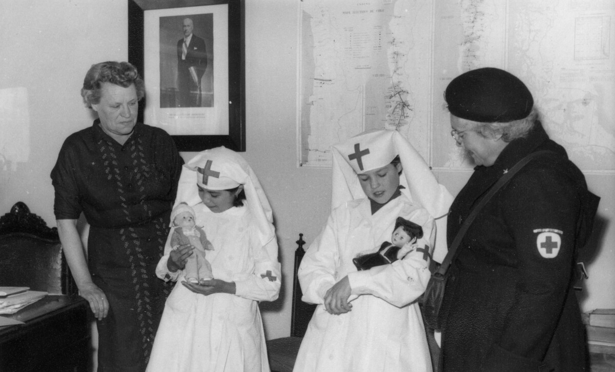 plano general de dos niñas de la cruz roja, mirando a las muñecas que tienen en sus brazos. Al costado izquierdo se encuentra Olga Boettcher, miembro de la institución y al lado derecho hay una mujer mayor también perteneciente a la cruz roja. Se encuentran en el interior de una infraestructura no identificada.