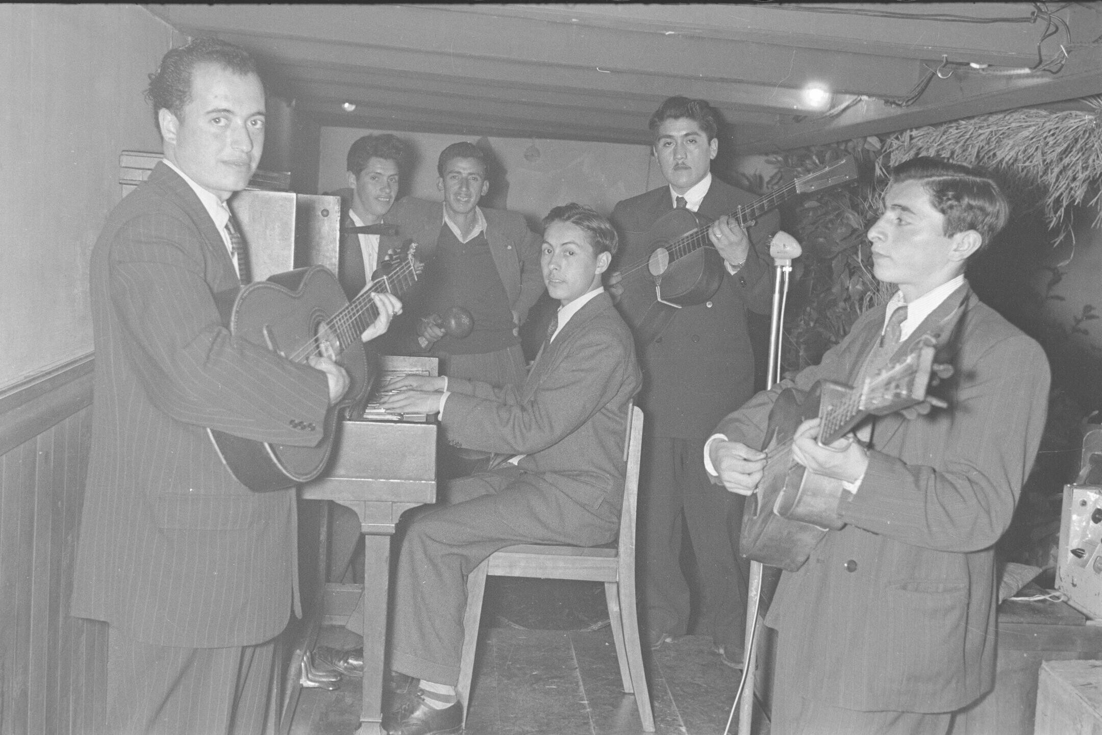 plano general de un grupo musical tocando sus instrumentos. Algunos de ellos están mirando hacia la cámara. Todos visten formalmente. Se encuentran tocando en el interior de un local no identificado, durante la celebración de una coronación a reina no identificada