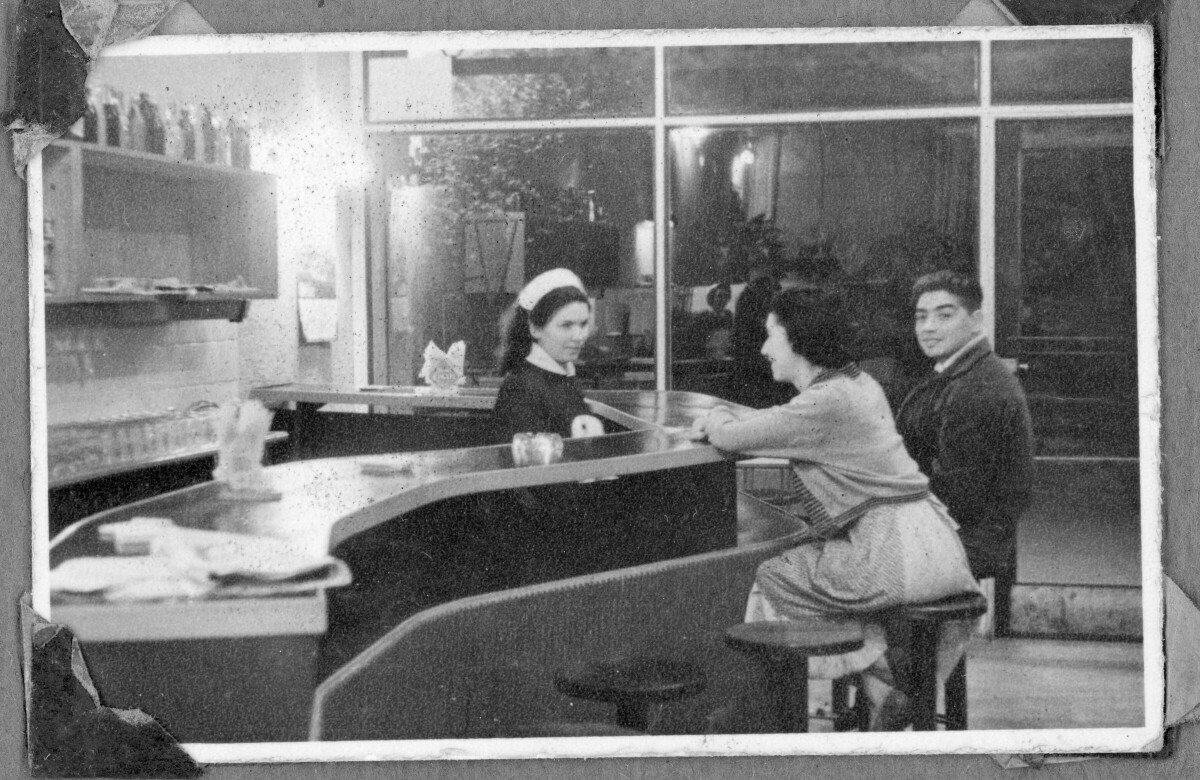 Vista interior de un restaurante o cafetería con tres personas dentro, dos de ellas sentadas en un banco en la barra.