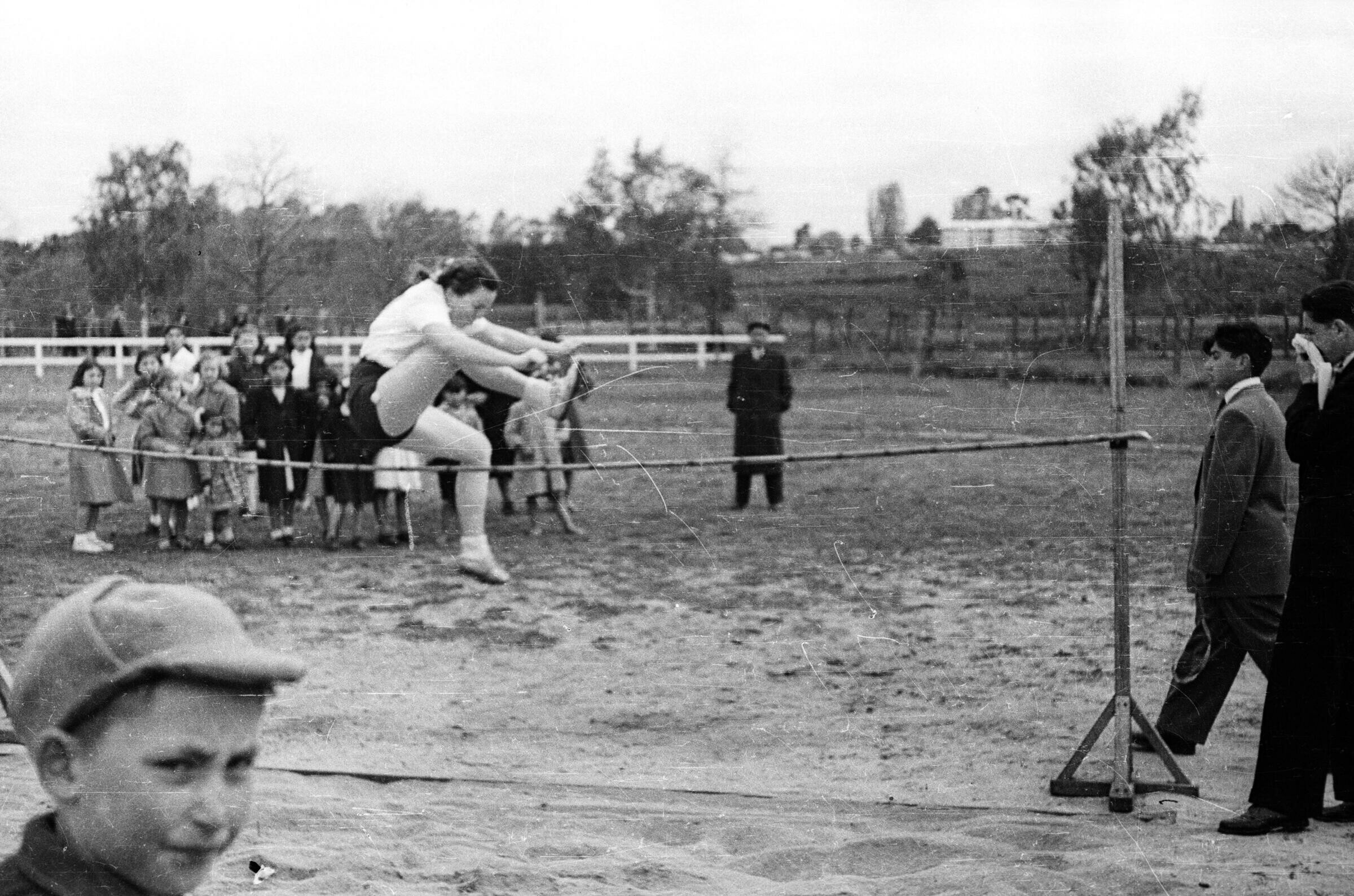 Plano entero de una mujer realizando un salto de altura, en el fondo hay un grupo de Mujeres y Niñas observándola. dos hombres jóvenes la observa mientras uno se limpia la nariz con un pañuelo y en el frente hay un niño con gorro mirando a la cámara. Están sobre una cancha de pasto.