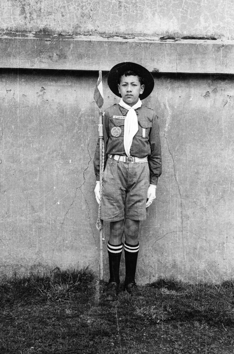 Retrato de plano entero de un niño posando de pie vestido de Scout, sosteniendo una vara con un banderín en la punta sobre el pasto en las afueras de una infraestructura