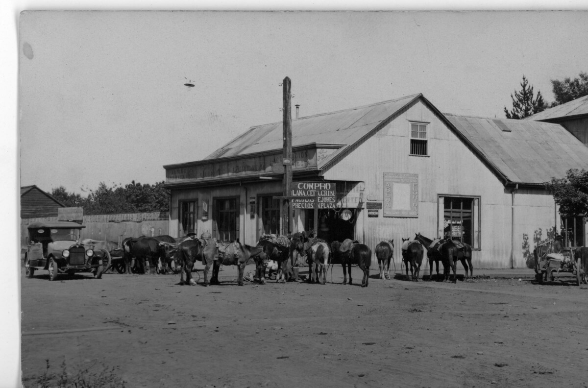 -plano general de una casa esquina utilizada como tienda. En sus afueras, tiene un letrero que dice --Compro lana, <ilegible> pago los mejores precios, plaza-- En sus afueras, hay un grupo de caballos con montura, parados esperando a sus dueños. A un costado hay un automóvil estacionado sobre la calle de tierra. El lugar corresponde a la ciudad de Loncoche-