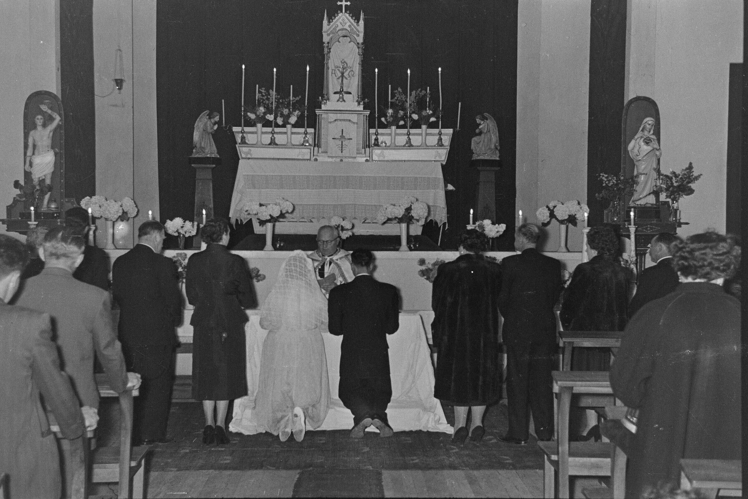 plano general de una pareja de novios, arrodillados en un reclinatorio. Ellos, al igual que sus padrinos de bodas y sus invitados, se encuentran de espaldas hacia la cámara. En el fondo, un sacerdote se encuentra llevando a cabo la misa de celebración de matrimonio en el interior de la parroquia San José