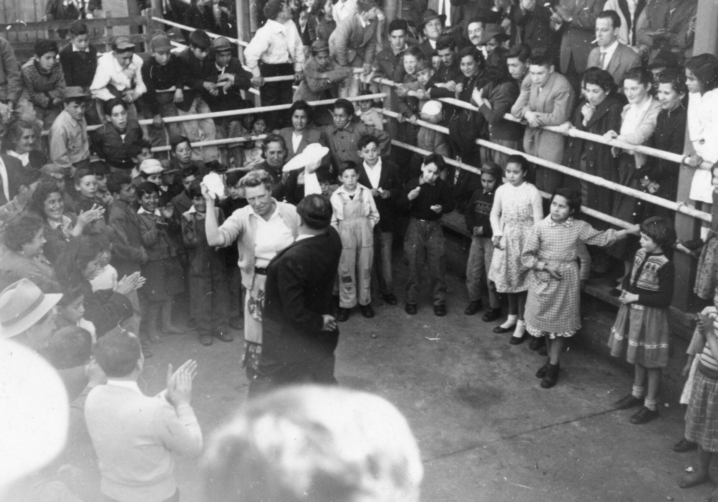 plano general de Olga Boettcher bailando un pie de cueca con un hombre vestido de Traje. Ambos agitan sus pañuelos mientras a su alrededor, un grupo de Mujeres, Niñas, Niños y Hombres aplauden como es costumbre, para avivar el baile. Se encuentran en un recinto no identificado