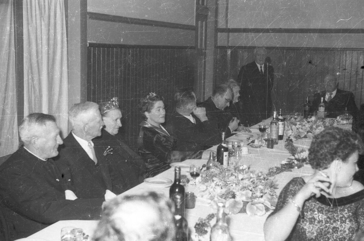 plano general de dos mujeres adultas con corona y grupo de Hombres con Trajes alrededor de una mesa con botellas de vino y copas, de pie al fondo un Hombre con micrófono, en lugar no identificado.