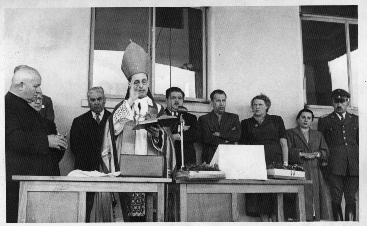 plano general de un obispo, leyendo la biblia frente a un micrófono. Le acompaña un sacerdote en un costado y un grupo de Hombres, Mujeres y un carabinero en el fondo. La primera mujer de izquierda a derecha es Olga Boettcher. Se encuentran en la inauguración del hospital Dr. Juan Morey de La Unión realizado en el frontis del edificio