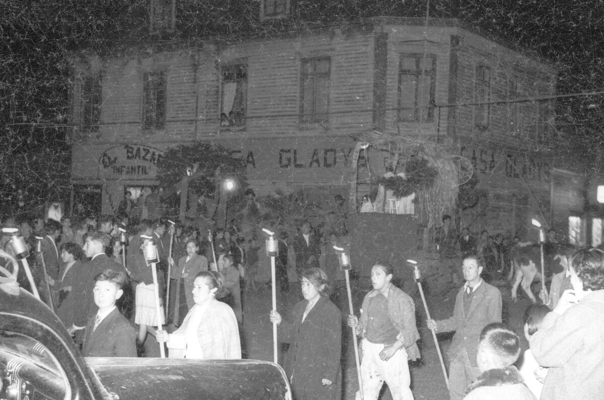 plano general de un grupo de Hombres y Mujeres caminando con una vela encendida sobre un palo. Están girando alrededor de un carro alegórico. Se encuentran en el frontis de la casa Gladys durante la noche