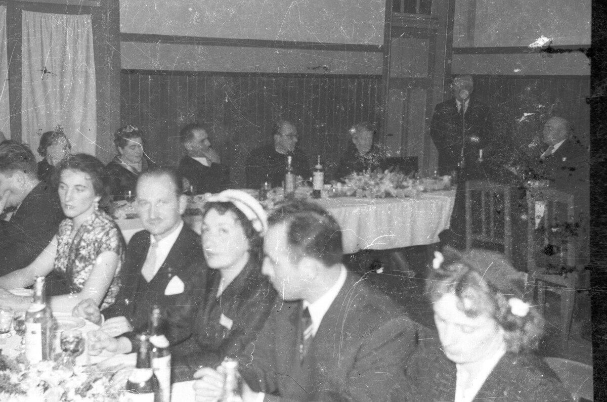 plano general de Hombres con Trajes y Mujeres con Vestidos, dos de ellas con una corona en la cabeza, sentados alrededor una mesa, al fondo un Hombre con un micrófono en un lugar no identificado