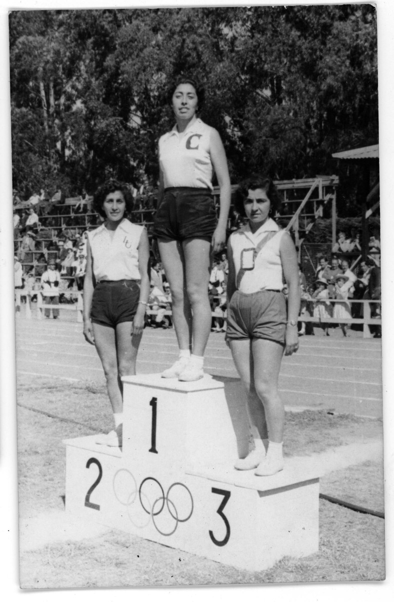 Plano general de tres atletas posando sobre una tarima de coronación del primer, segundo y tercer lugar. En el segundo lugar se encuentra Raquel Latorre. Se encuentran en un estadio no identificado, durante un zonal sur de atletismo en Tomé