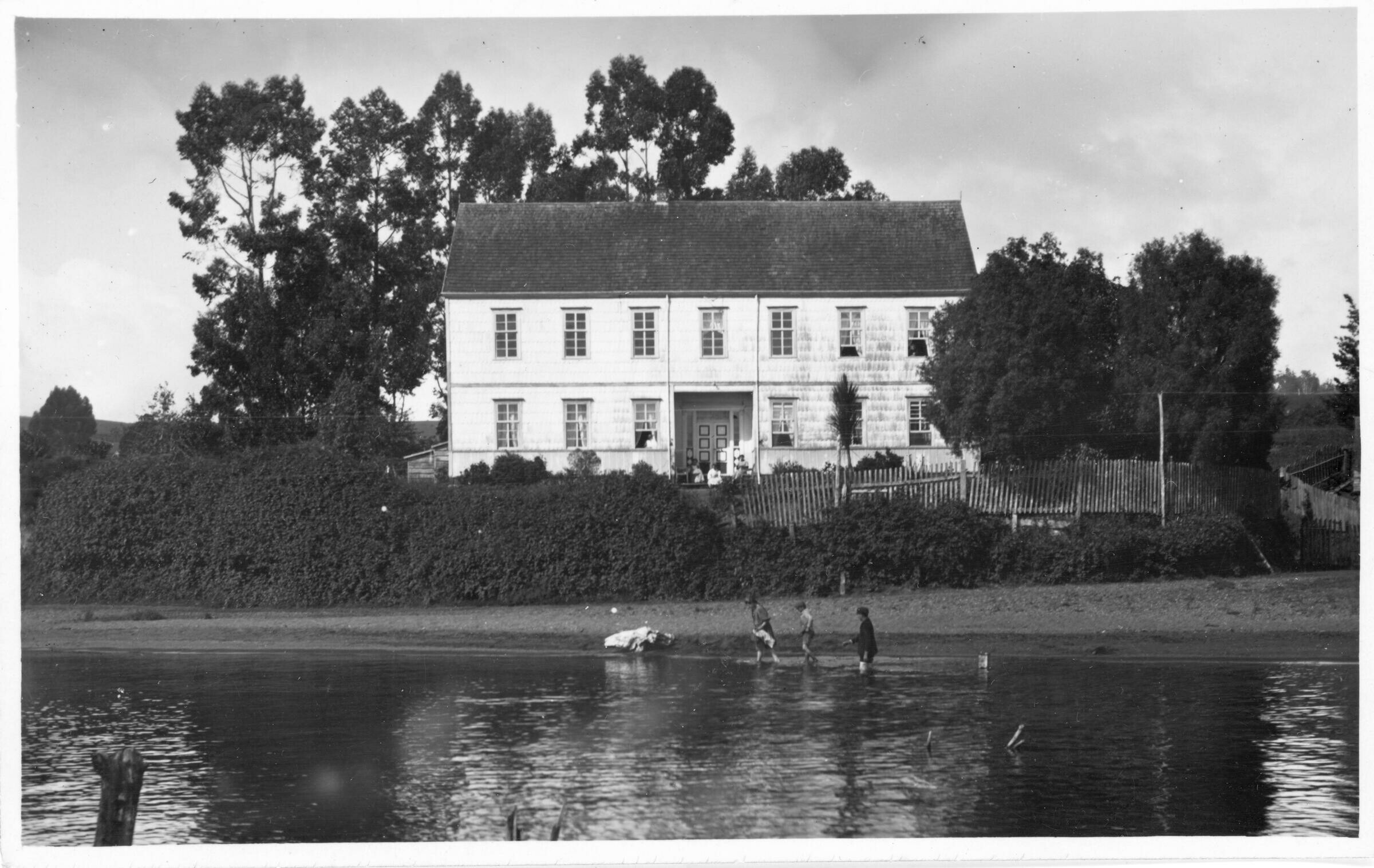 plano general de tres niños caminando en el agua en el frontis de una casona de dos pisos, mientras que en la entrada de la estructura, hay tres personas sentadas. La casa queda a orillas de un RÍO no identificado