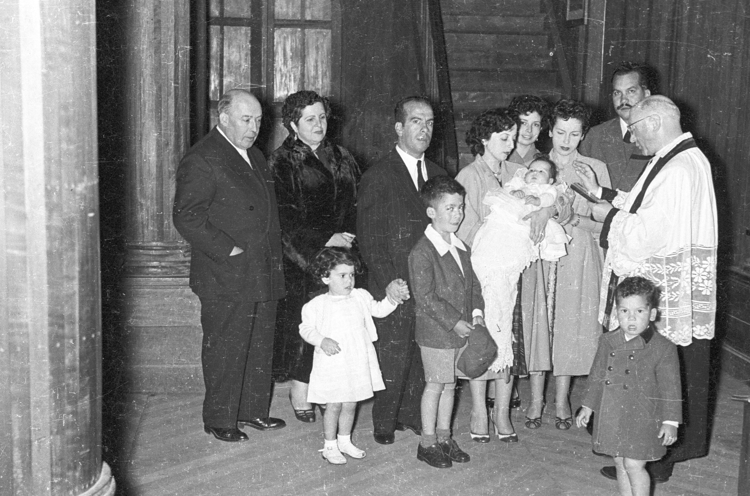 Plano entero de un sacerdote, leyendo la biblia frente a una bebé la cual está siendo cargada por una mujer. A su alrededor los acompaña un grupo de Hombres, Mujeres, dos niños y un niña mientras se lleva a cabo su bautizo. Están en el interior de la parroquia San José