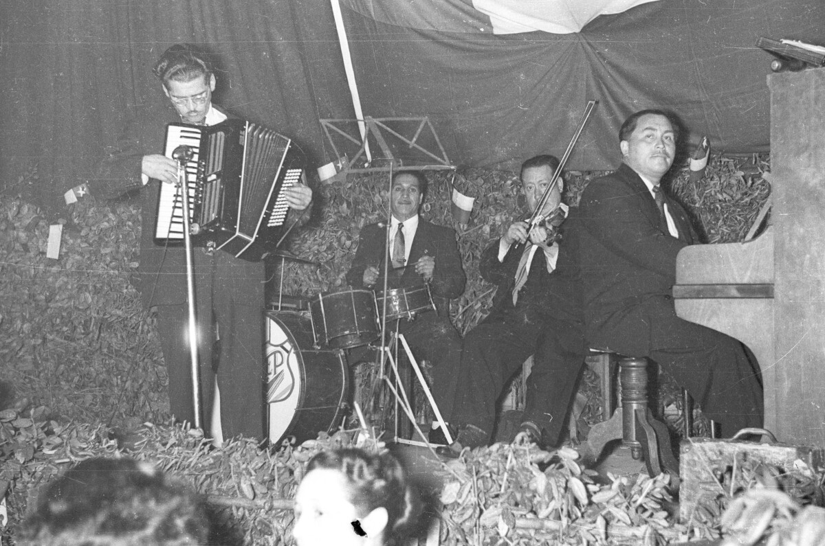 Plano entero de un grupo musical no identificado, realizando su presentación sobre un escenario. Uno de los hombres se encuentra tocando un acordeón, de pie junto al micrófono. Están en el interior de un local durante una celebración de fiestas patrias.