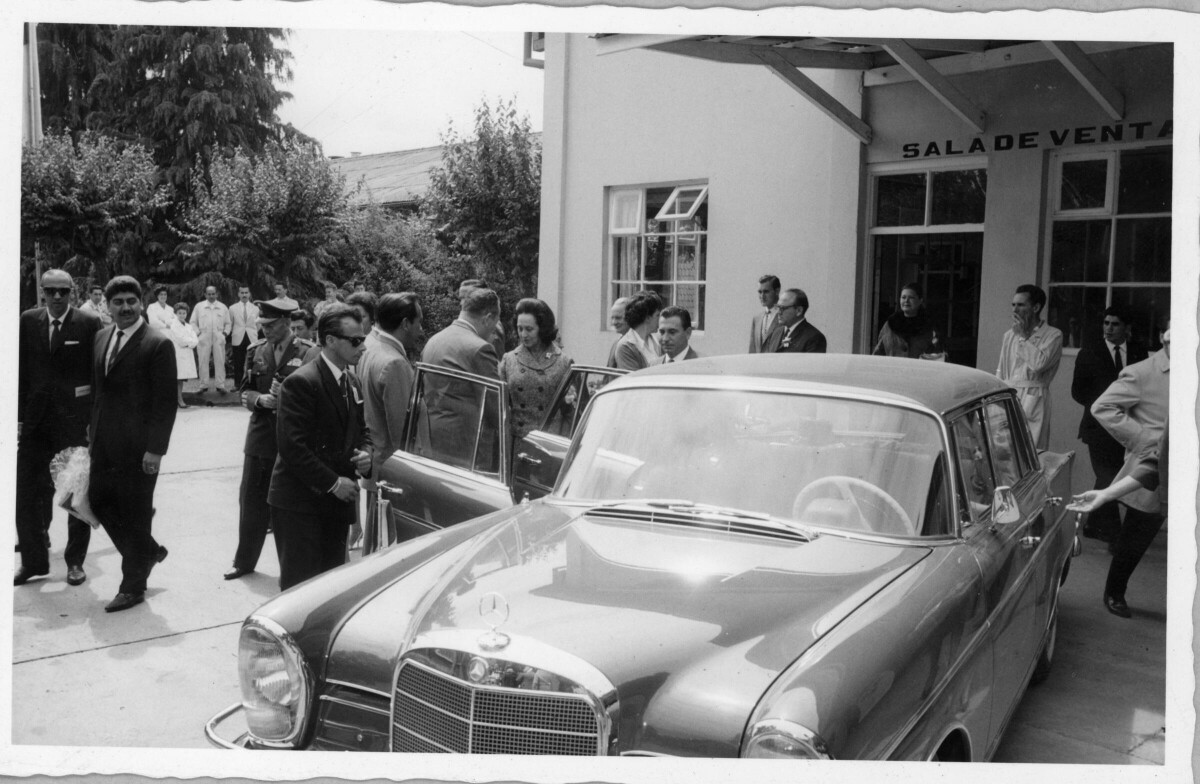 Plano entero de un hombre conversando con Eva Sámano a costado de un auto durante su visita a La Unión. A su alrededor, hay otro grupo de hombres y mujeres caminando en las afueras de la sala de ventas del estadio Carlos Vogel, durante un homenaje realizado en su honor