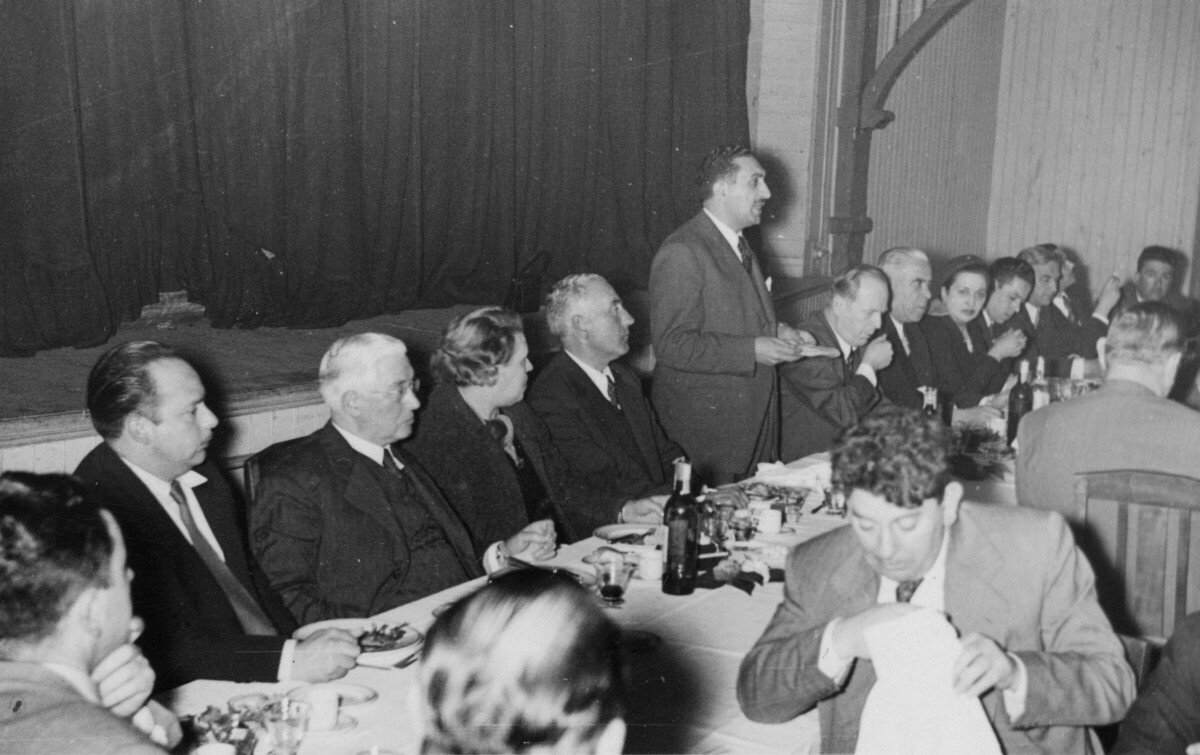 plano general de un grupo de personas sentadas alrededor e una larga mesa mientras en el medio hay un hombre de pie, dirigiéndose a ellos. La tercera mujer sentada de izquierda a derecha es Olga Boettcher, primera gobernadora de La Unión. Se encuentran en una infraestructura no identificada durante un banquete