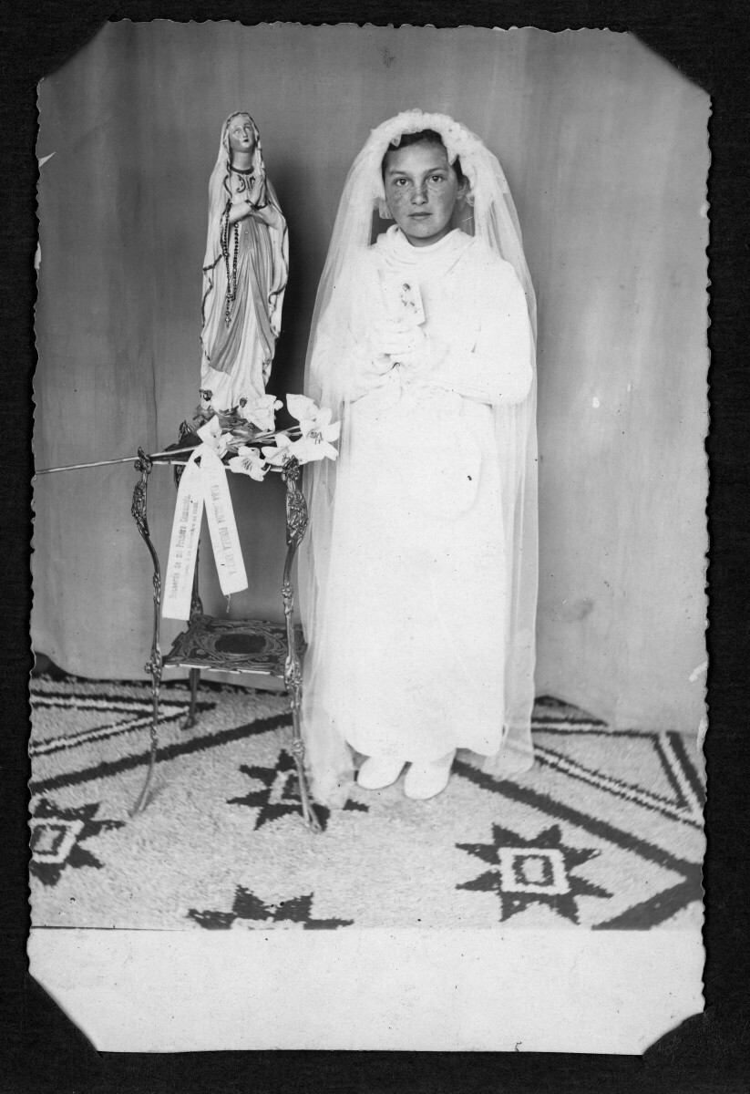 Retrato de estudio de Plano entero de una niña posando de frente parada junto a una virgen de yeso. Lleva puesta la ropa típica de la época utilizada para las primeras comuniones