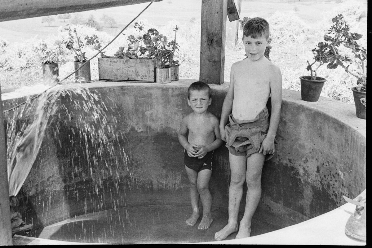 Plano entero de dos niños descalzos y con el torso desnudo, bajo el chorro de una llave de riego, ubicada en el interior de un pozo. En los bordes hay distintas plantas en maceteros. El pozo se encuentra en el antejardín de una casa de un piso