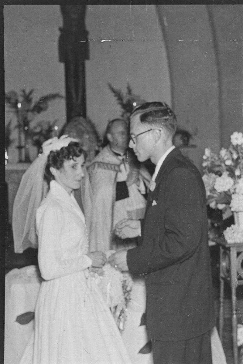 Plano medio largo de un novio acercándose a la novia. En el fondo, hay un sacerdote parado con las manos cruzadas. Están en el altar durante la misa de celebración de su matrimonio, realizada en el interior de la parroquia San José