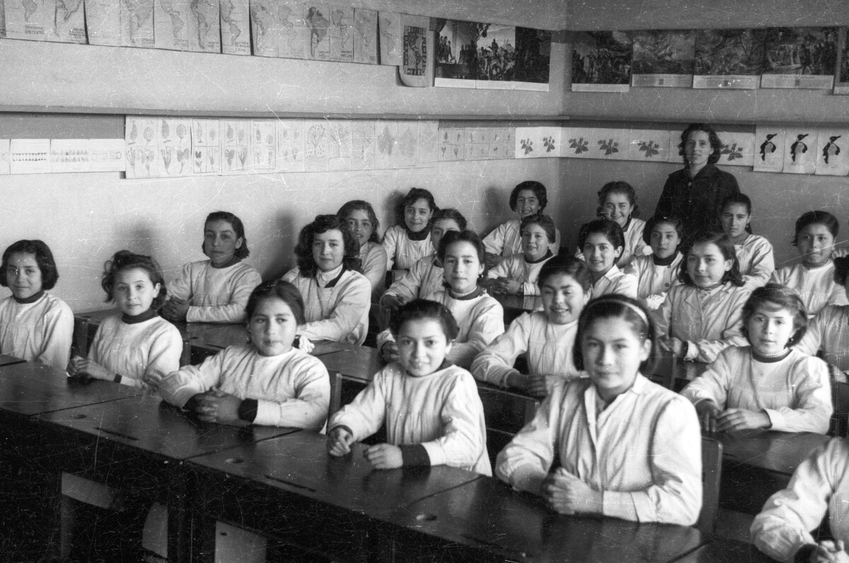 Retrato grupal de plano entero de niñas posando sentadas frente a sus pupitres y una profesora en el fondo de la sala de la escuela superior de niñas Nº2