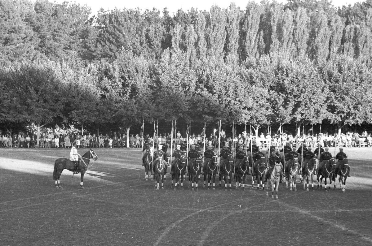 plano general de un carabinero montado a caballo junto a un grupo de jinetes, quienes se encuentran formados en hileras sosteniendo una valla en la mano mientras realizan su presentación. En el fondo, Niñas, Niños, Hombres y Mujer los observan sentados a un costado de la cancha ubicada en el estadio Carlos Vogel